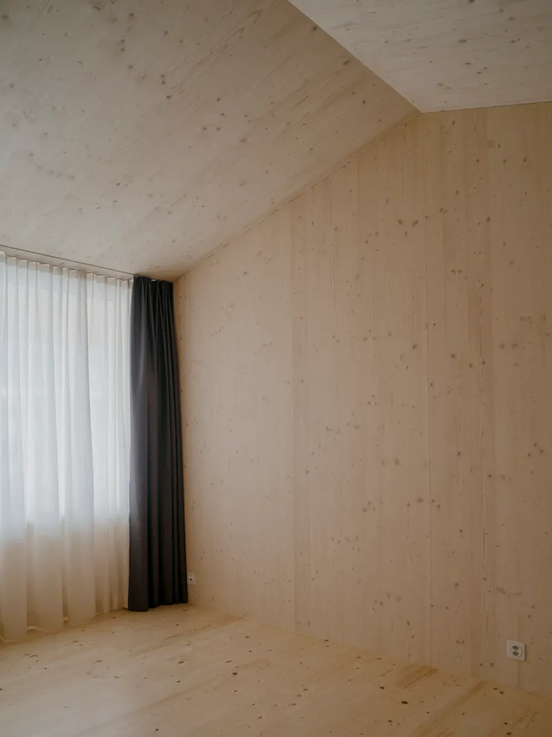 Interior corner of pale timber-lined room with sloped ceiling and layered white and grey curtains