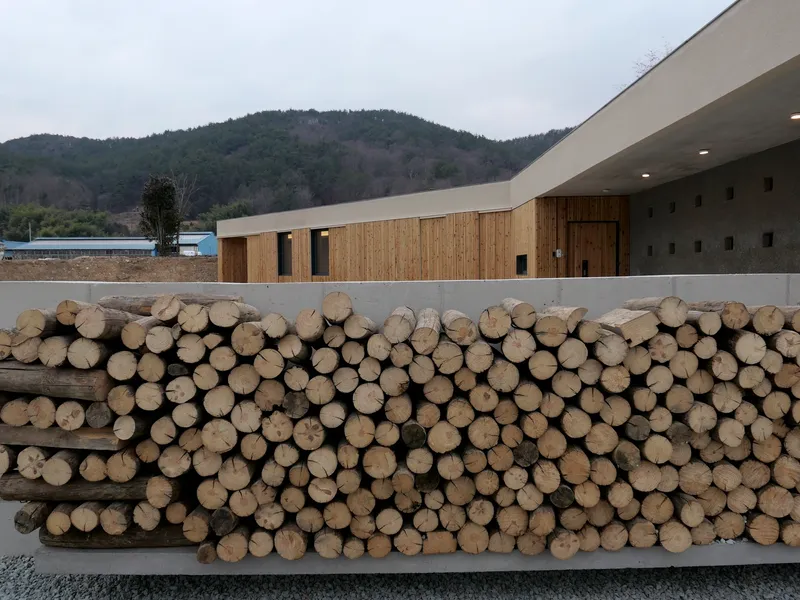 Stacked firewood logs stored in concrete planter alongside timber-clad volumes and hillside backdrop