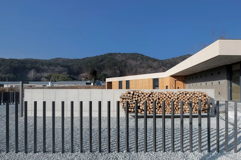 Vertical slat fence and stacked firewood storage in front of timber-clad volumes under a clear blue sky