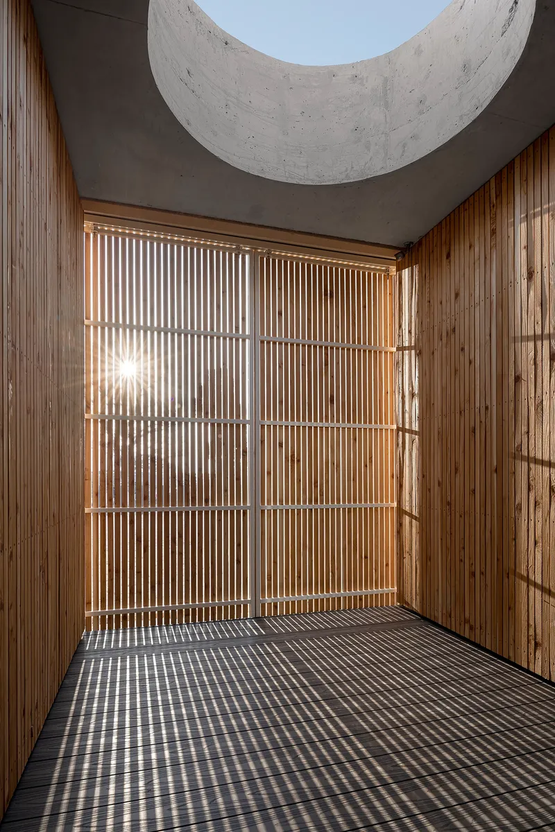 Timber slat screen and concrete ceiling with circular skylight opening casting striped shadows across the floor