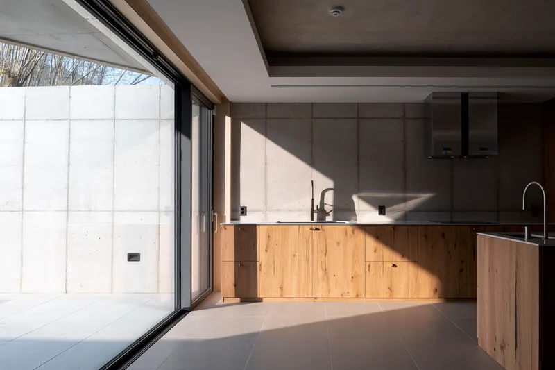 Kitchen with timber cabinetry and concrete walls beneath exposed ceiling beams with afternoon sunlight streaming through glazing