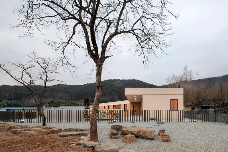 White concrete house behind vertical slatted fence with bare winter tree and hillside backdrop