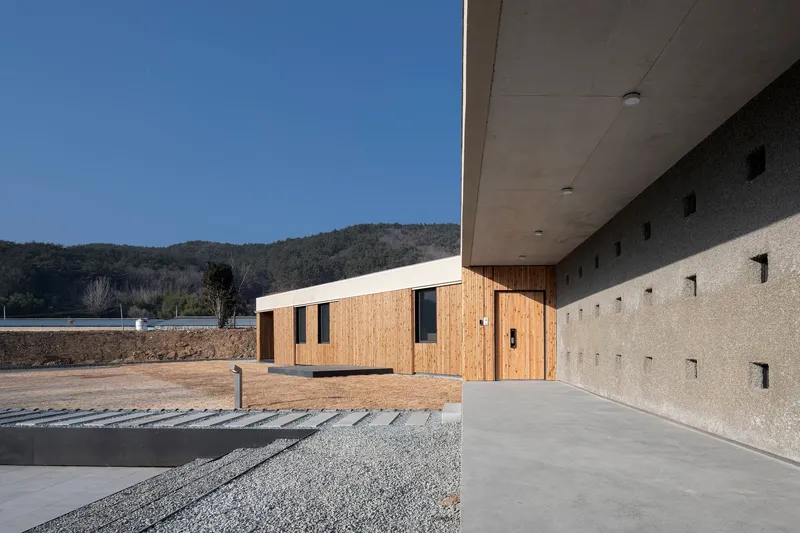 Covered walkway with concrete soffit beside gravel court and timber-clad wing under clear sky