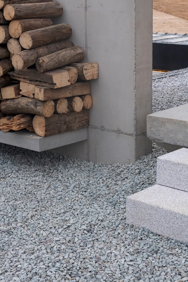 Stacked firewood against concrete column base with stone steps and gravel surround