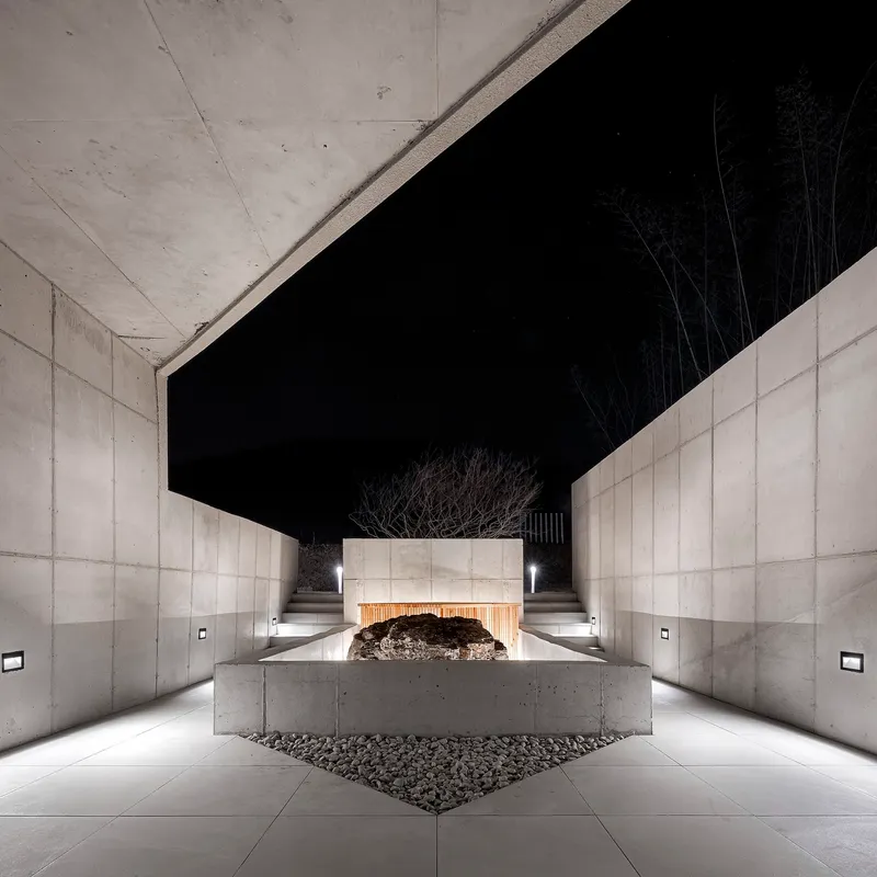 Covered entrance passage with concrete walls, overhead beams and a central planted tree at night