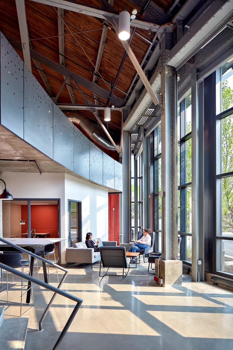 Lounge seating area beneath exposed timber trusses and metal ductwork with afternoon sunlight streaming through tall windows