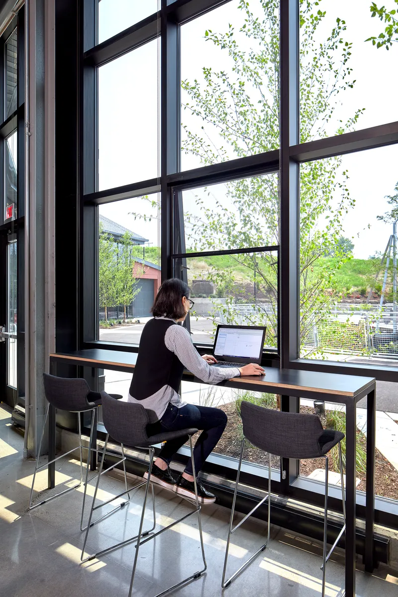 Person working at a standing-height table beside floor-to-ceiling windows overlooking exterior trees and parking lot