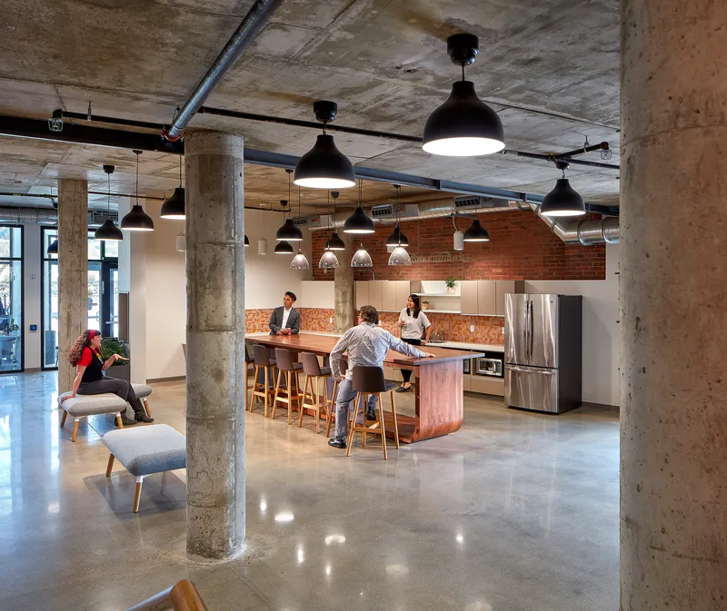 Open communal kitchen with exposed concrete columns and pendant lights as people gather at the central island