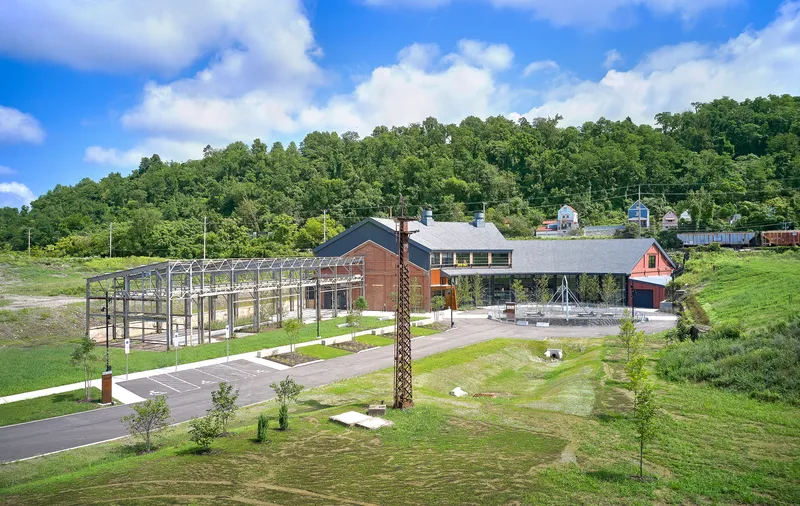 Site view showing the main building with weathered steel panels, glass facades and an open metal frame structure on the left