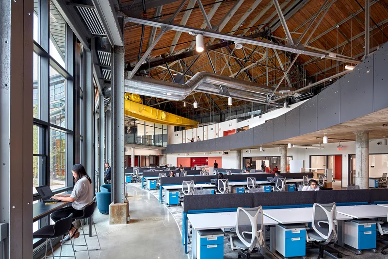 Double-height workspace with exposed wood bowstring trusses, concrete columns and rows of open office workstations below a mezzanine