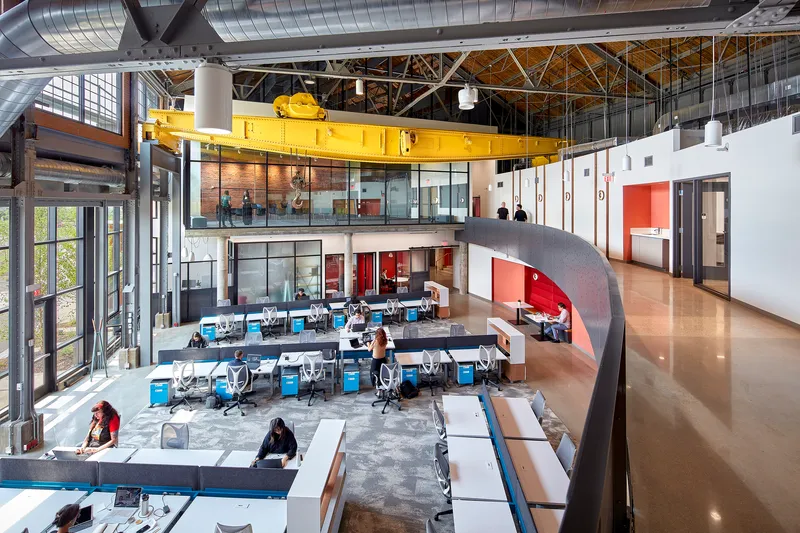 Interior view showing the yellow overhead crane beam, curved mezzanine ramp and open work floor with desk clusters