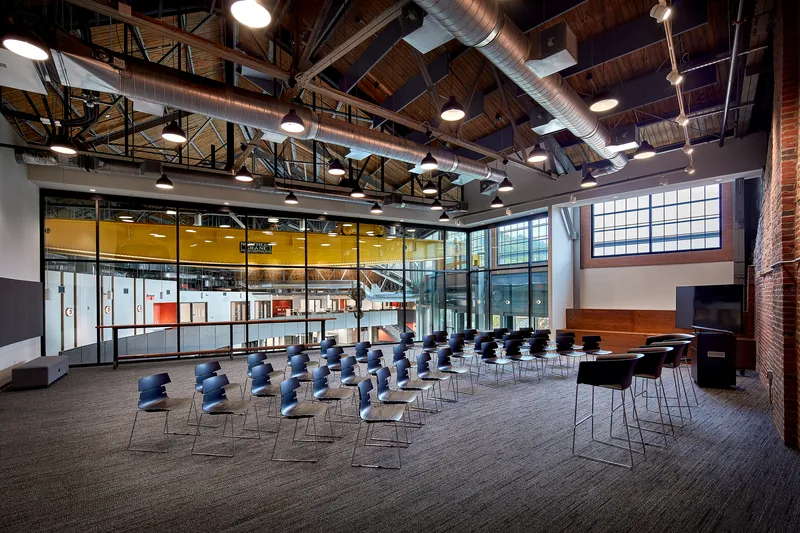 Double-height multipurpose hall with exposed timber roof trusses and rows of blue chairs facing interior windows
