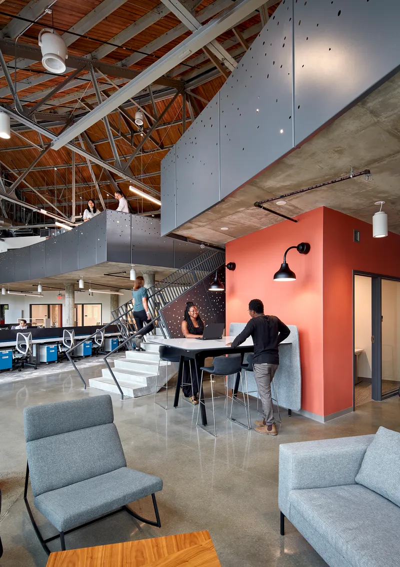 Open workspace with coral-colored partitions beneath white steel trusses and timber ceiling with three people conversing