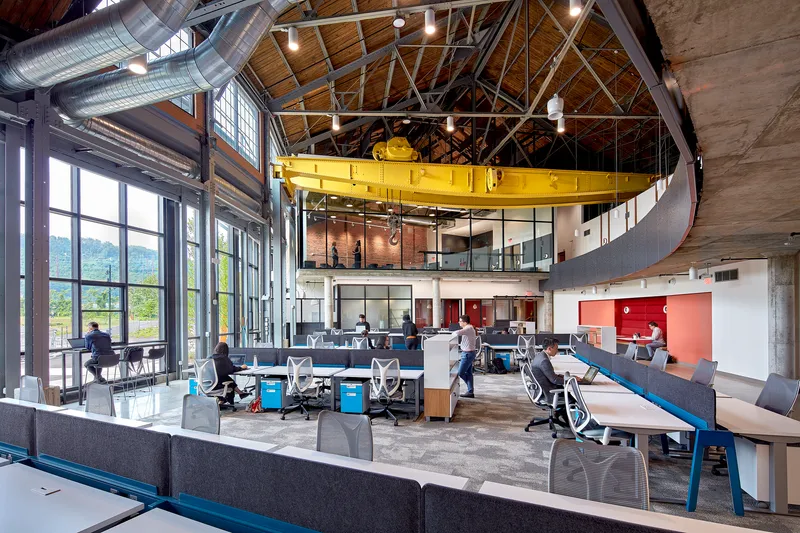 Multi-level interior with yellow suspended crane structure above workstations and conference rooms under exposed timber trusses