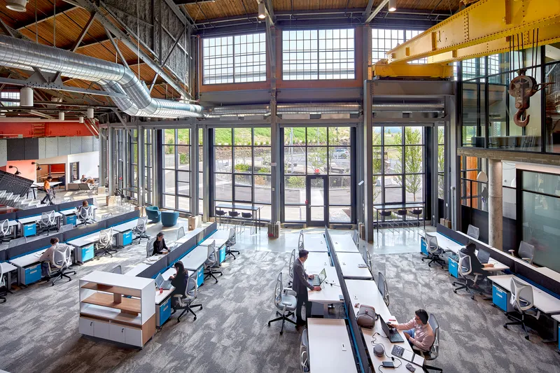 Double-height workspace with exposed timber roof trusses, steel columns, and rows of desks below clerestory windows