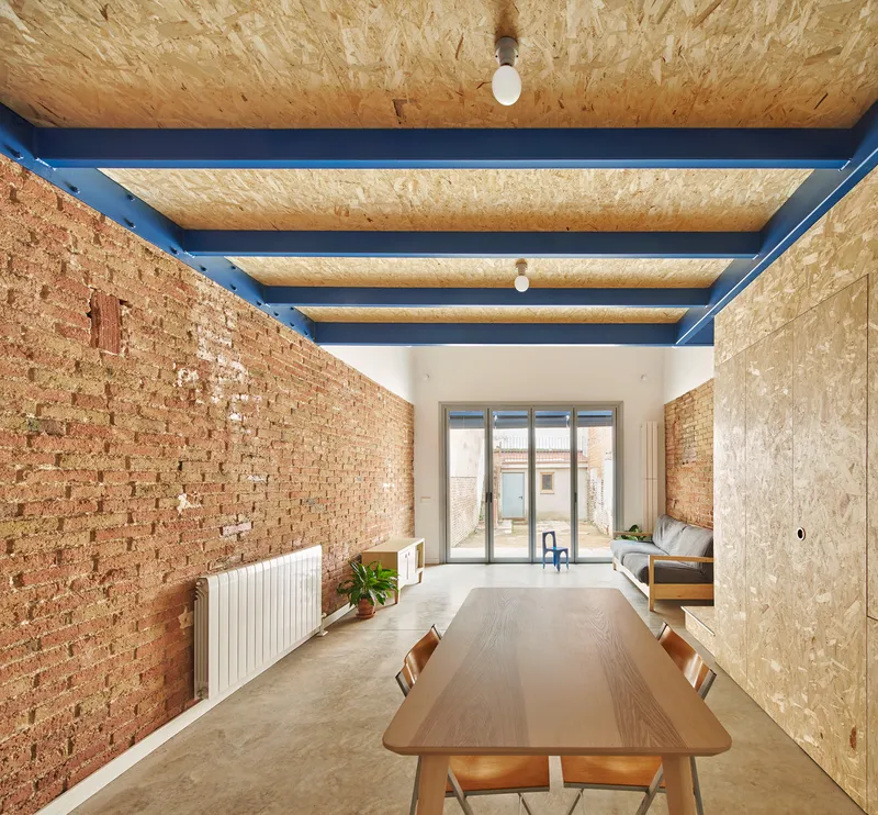 Dining table beneath blue painted steel beams with oriented strand board ceiling panels and brick walls