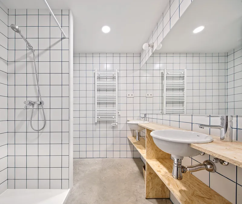 Bathroom with white square tile walls and plywood vanity counter supporting two basin sinks