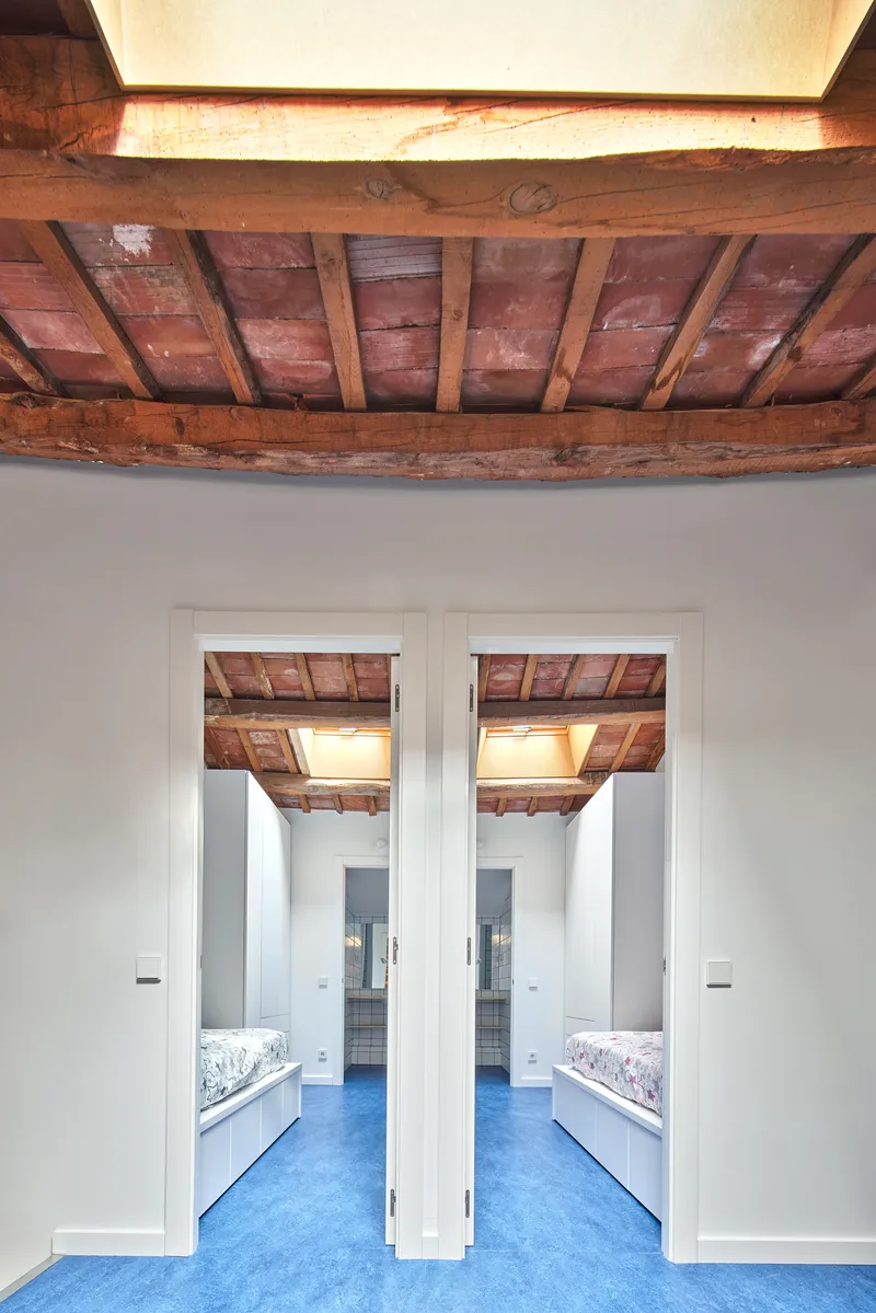 View through twin doorways into mirrored bedrooms with blue rubber flooring and restored timber beam ceilings