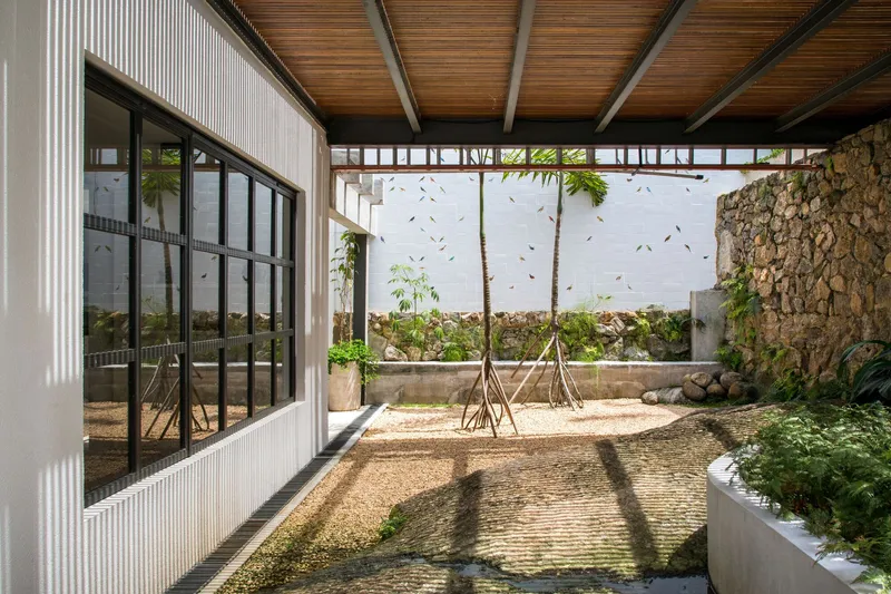 Covered courtyard with gravel floor, young trees, stone wall and timber ceiling casting afternoon shadows