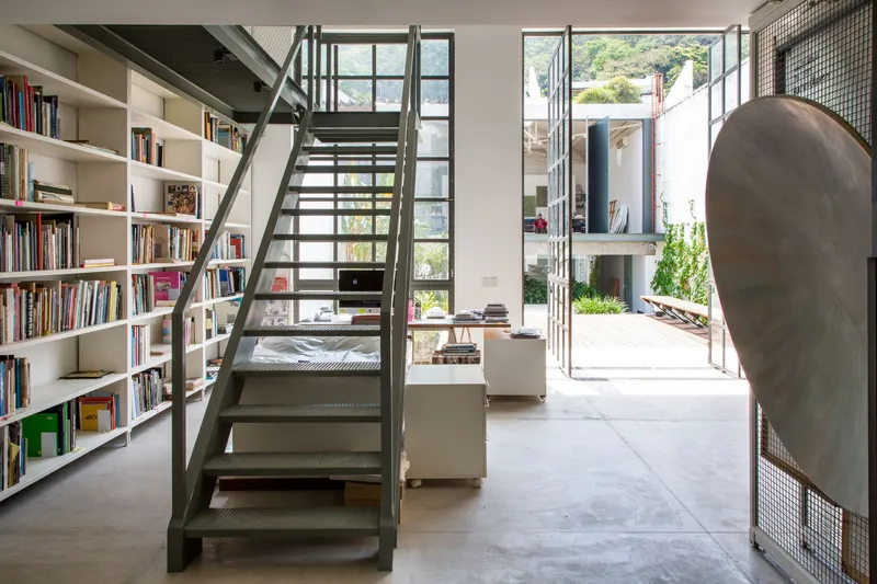 Steel staircase with perforated treads rising beside a floor-to-ceiling bookshelf in a double-height living space