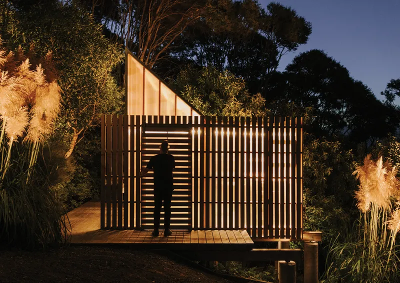 Illuminated timber slat screen with figure silhouette at dusk surrounded by grasses and trees
