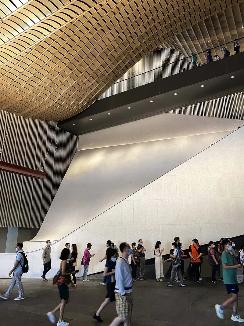 Curved wood-paneled ceiling above a sloping white wall as visitors queue in the foyer