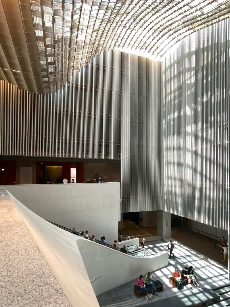 Multi-level atrium with vertical metal louvers filtering daylight onto white concrete balconies