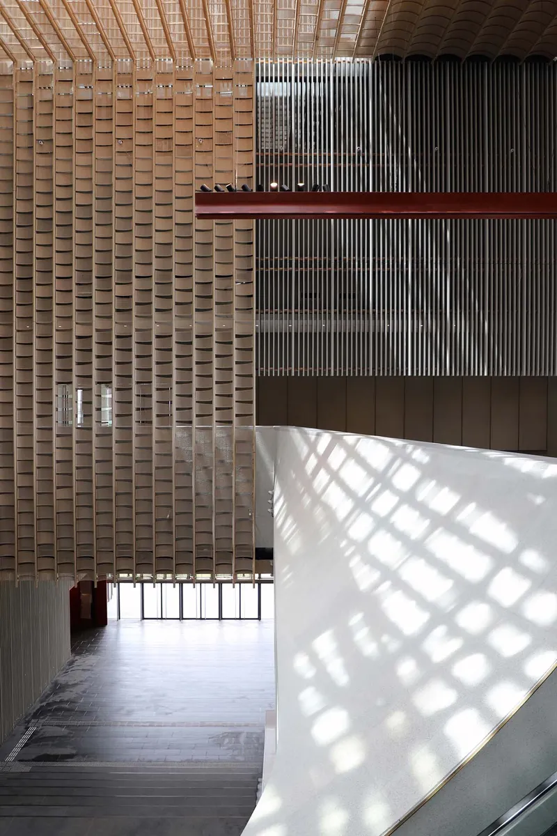 Multi-story lobby with woven timber ceiling and vertical slat screen filtering daylight onto the white wall