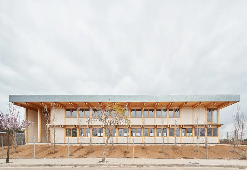 Two-story timber-framed facade with deep overhanging roof and newly planted trees in bare earth