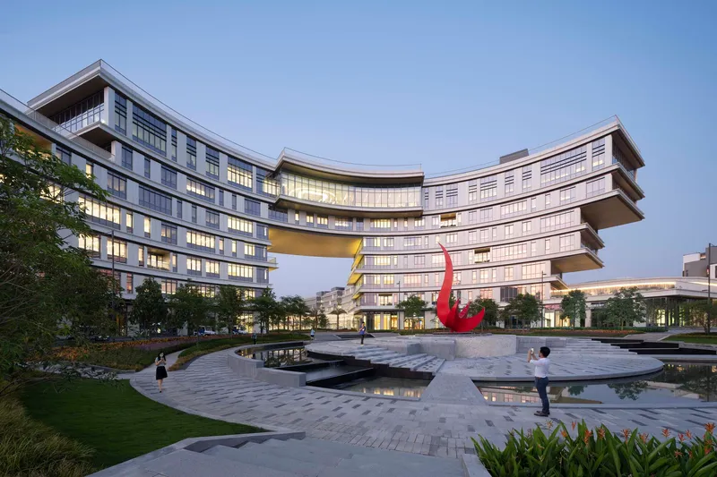 Courtyard with reflecting pools and red sculpture beneath a curving skybridge connecting two multi-story wings at dusk