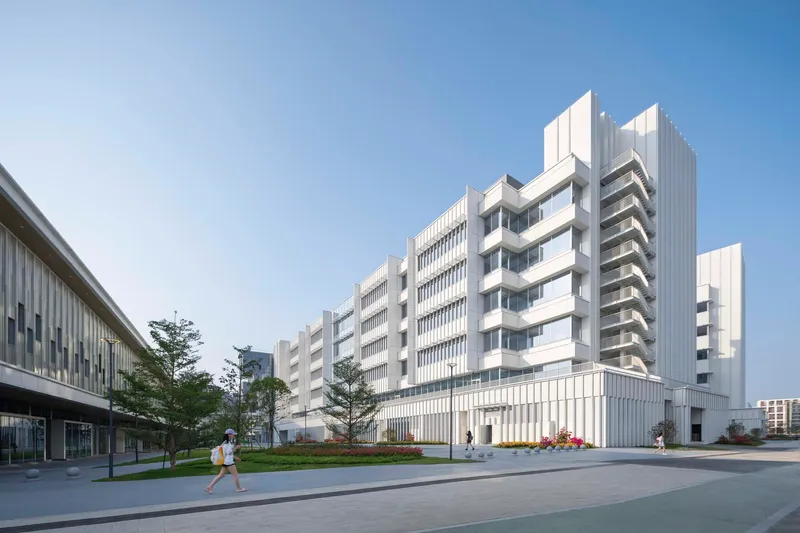 White vertically ribbed facade with stepped massing and a lone pedestrian walking along the tree-lined plaza