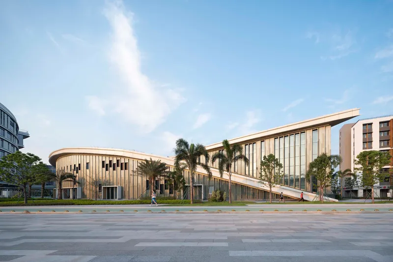 Curved two-story pavilion with vertical metal fins fronting a vast paved plaza lined with palm trees