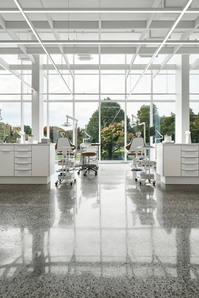 Interior workspace with dental chairs and equipment facing floor-to-ceiling windows overlooking trees