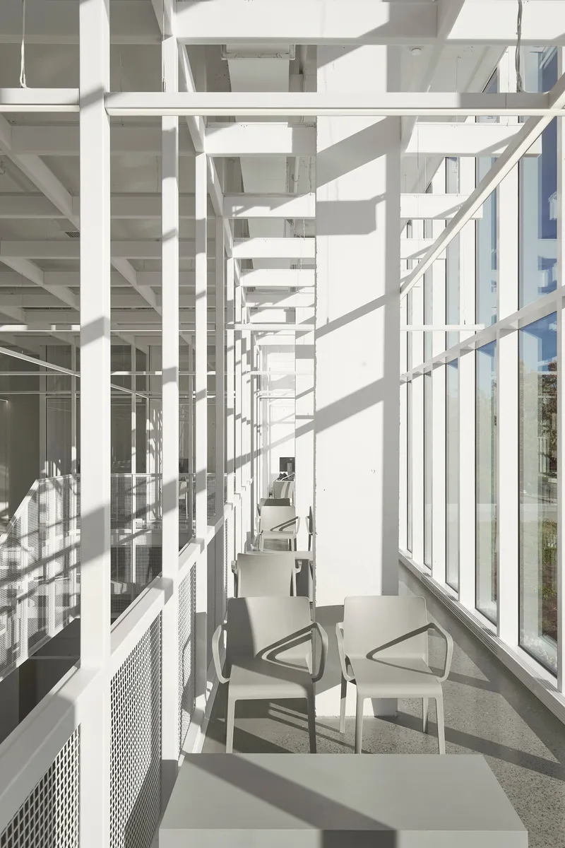 Narrow corridor along the glazed facade with perforated metal balustrade casting sharp shadows on the terrazzo floor