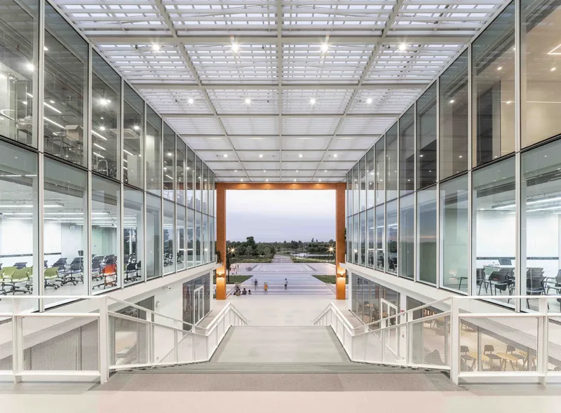 Glass-walled atrium bridge with gridded translucent ceiling panels overlooking an outdoor courtyard at twilight