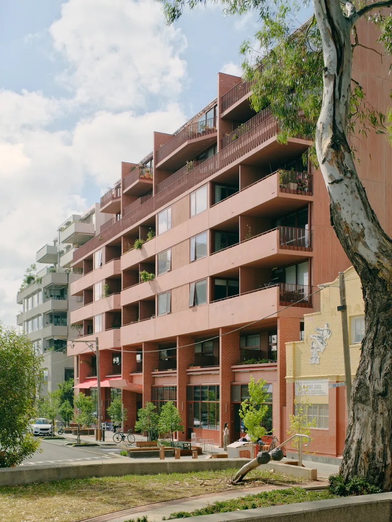 Stepped terracotta facade with planted balconies viewed from tree-lined street under partly cloudy sky