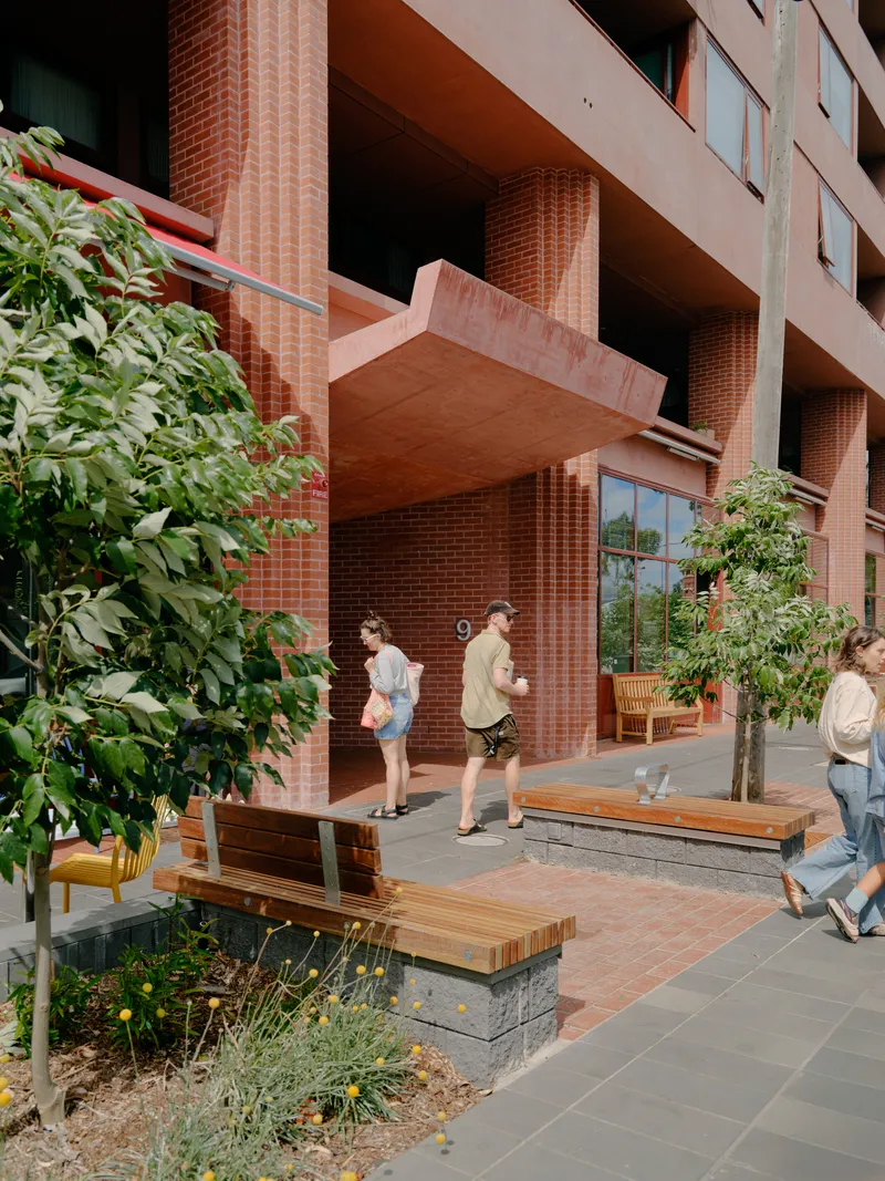 Timber bench seating beside planted beds at the base of red brick ground floor columns