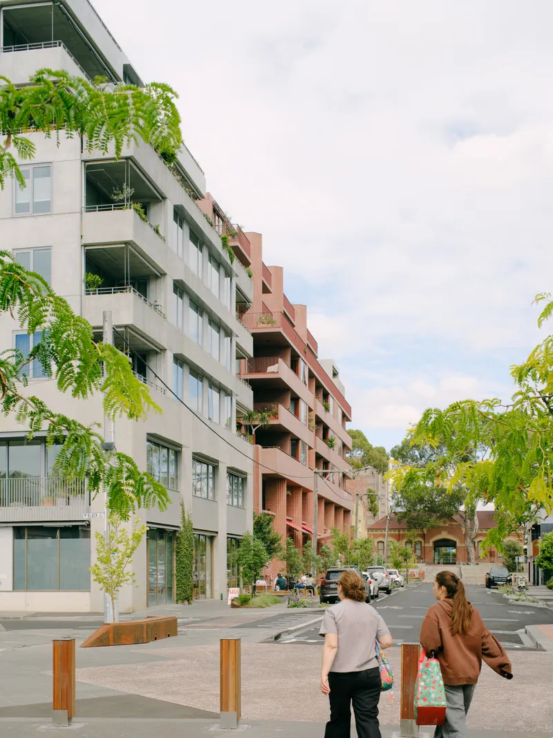 Street view of stepped residential volumes with planted balconies and two pedestrians crossing the intersection
