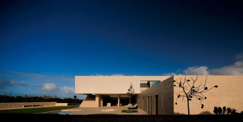 Side elevation of the stucco volume with recessed colonnade and young tree at dusk