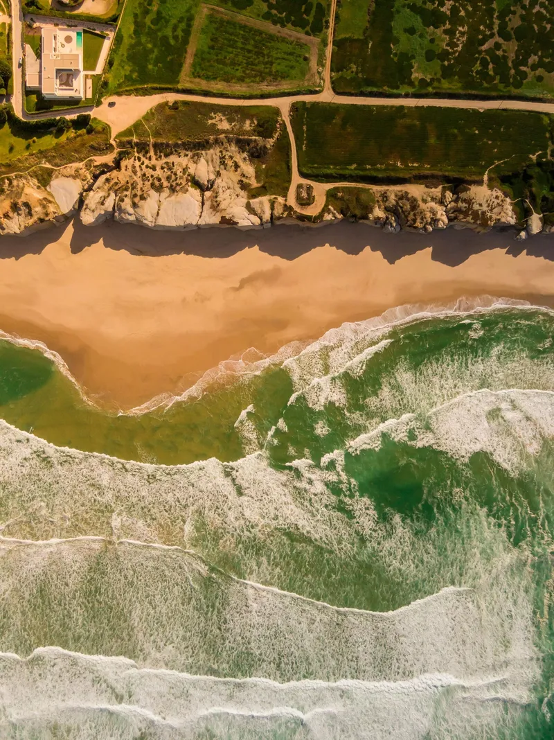 Aerial view of white sand beach meeting green ocean waves with a coastal building visible inland