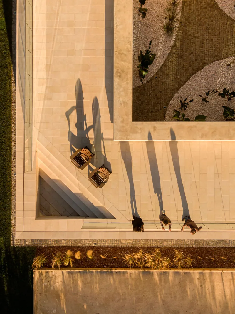 Overhead view of pale stone terrace with two wicker lounge chairs and three people casting elongated shadows