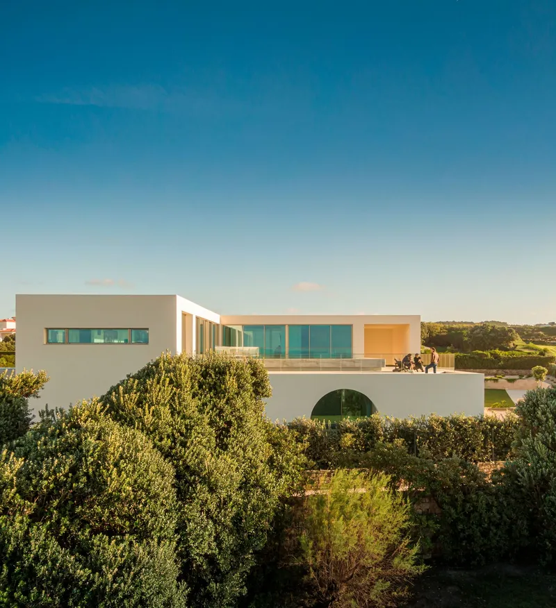 Two-story white residence with horizontal ribbon windows and arched opening at base surrounded by dense vegetation