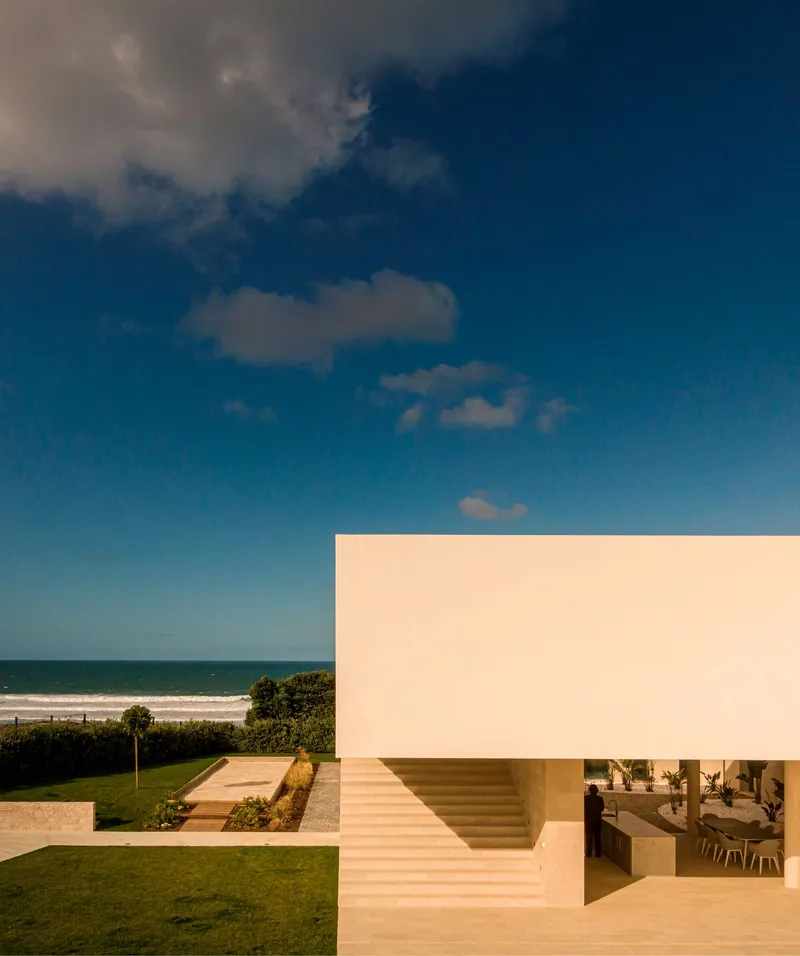 Corner view of white rendered facade with recessed staircase and ocean backdrop under blue sky
