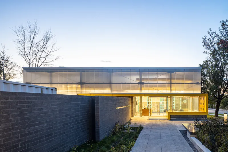 Translucent polycarbonate volume hovering above dark brick walls and yellow-framed entrance at dusk