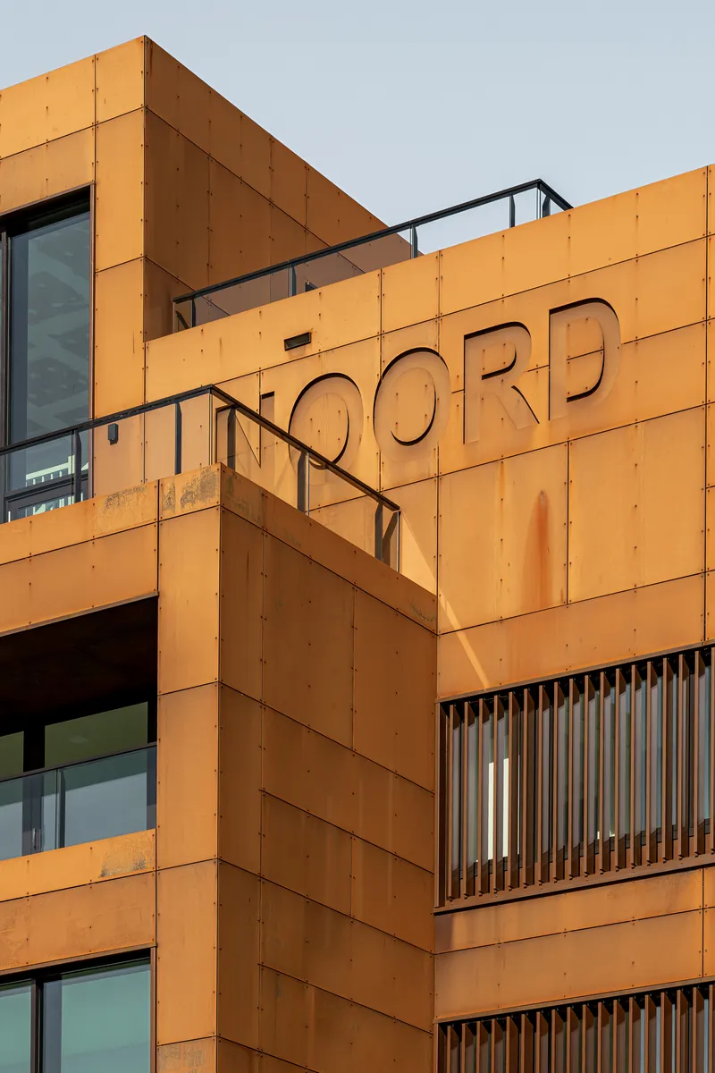 Close-up of orange-cladded facade with cut-out lettering and vertical slat screening at dusk