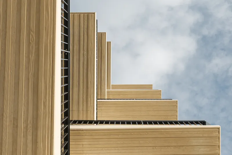 Stepped timber-clad facade volumes with vertical slat screens under a partly cloudy sky
