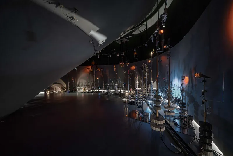 Exhibition gallery with reflective floor displaying illuminated glass sculptures along dark curved walls