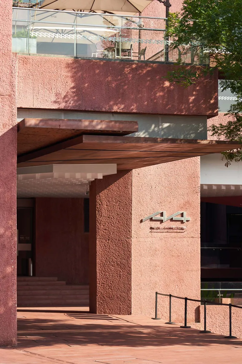 Layered facade with projecting timber roof plane and textured red walls beneath dappled tree shadows