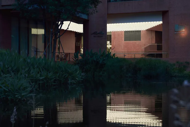 Evening view of illuminated interior volumes reflected in pond with lush foreground vegetation
