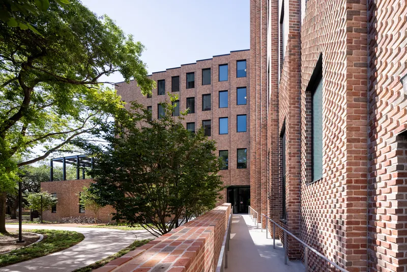 Patterned brick wall with vertical slits beside a shaded pathway and mature tree